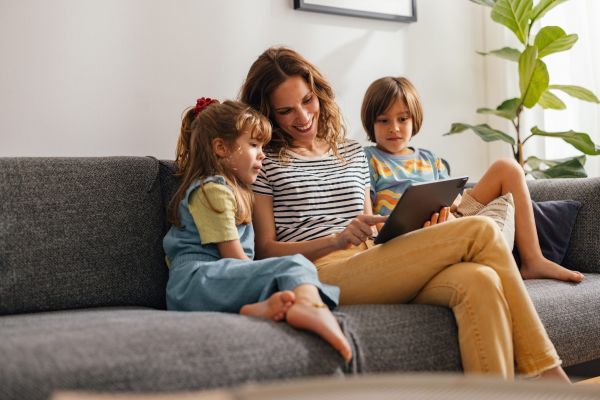 A woman and two children sit on a sofa, smiling while looking at a tablet.