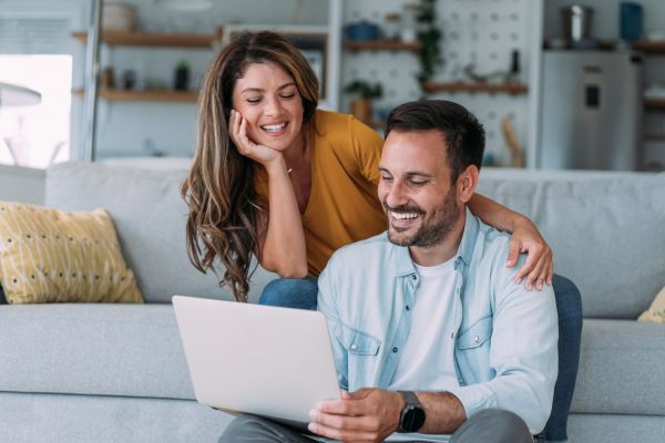 A smiling couple sits on a couch, looking at a laptop together in a cozy living room.