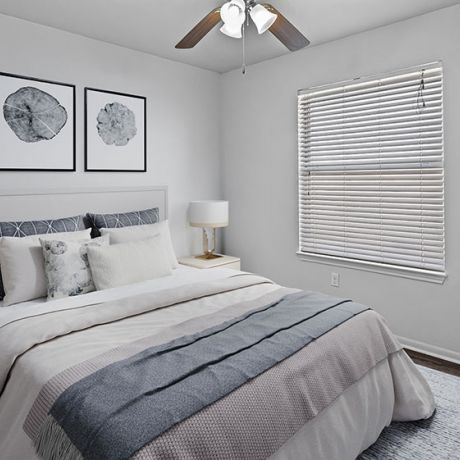 A cozy bedroom at Encino Park Apartments with a neatly made bed, two lamps, wall art, a plant, and a window with blinds.