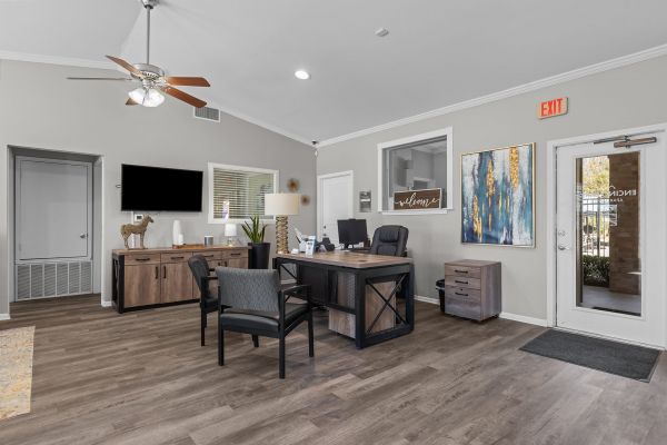 Modern business center at Encino Park Apartments with wood flooring, desk, chairs, artwork, and decor. Large windows and a wall-mounted TV.